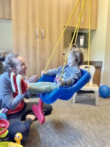 child sitting in a swing with a therapist seated on floor beside her